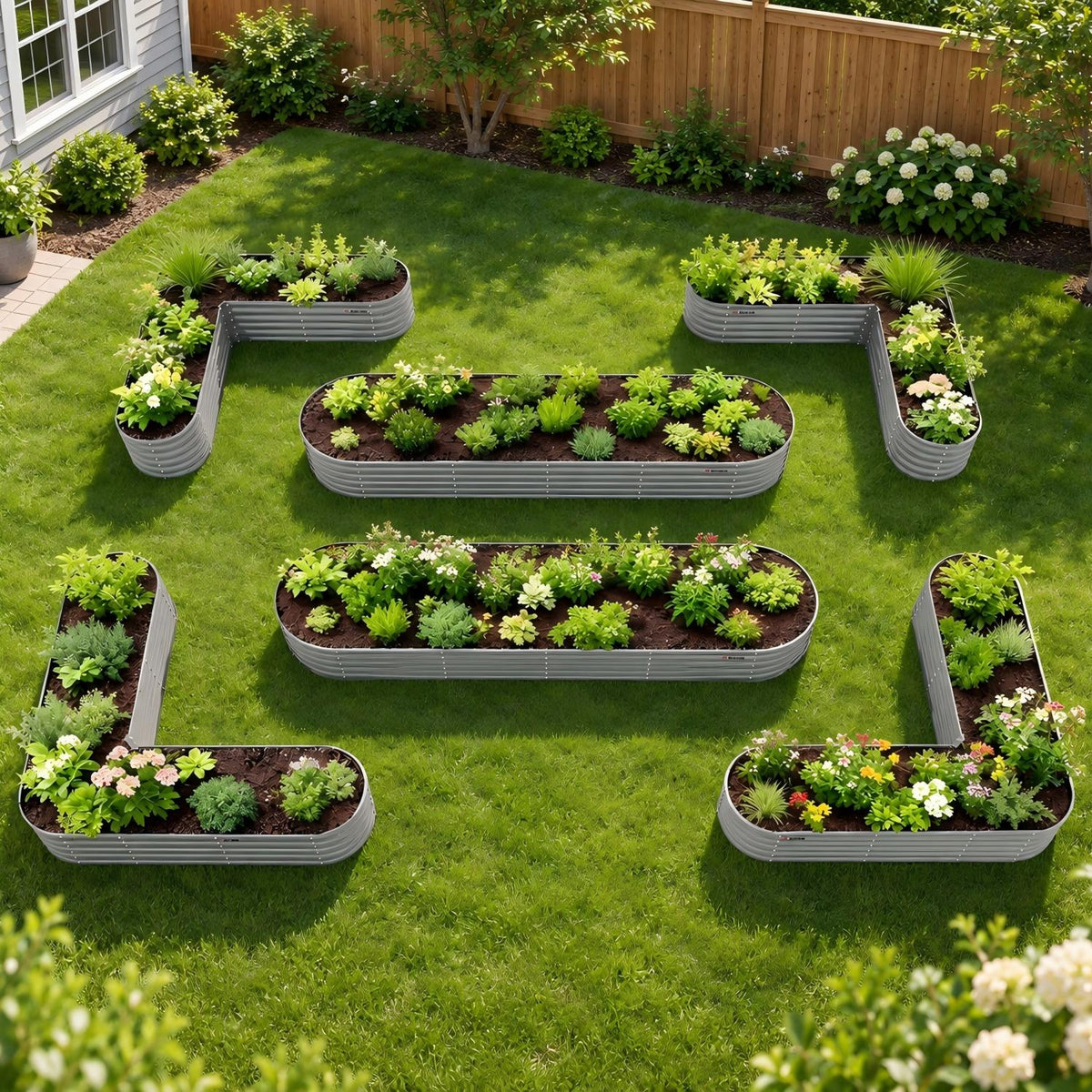 Six galvanized steel L-shaped raised garden beds arranged on a green lawn in a backyard setting, filled with various green plants and small flowers. 