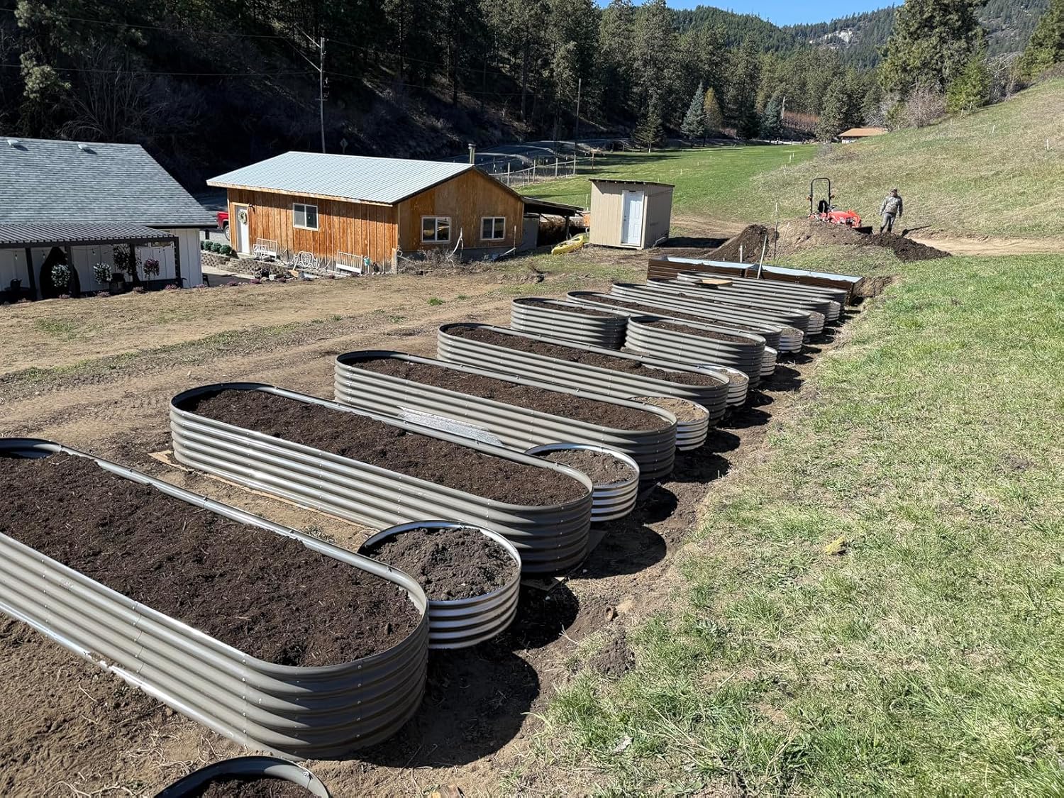 Row of metal garden beds on a farm with buildings and trees in the background 